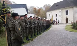 Eine Reihe von Soldaten in grünen Uniformen und Mützen steht in Formation vor einem Gebäude mit Flaggen und Menschen in der Nähe.