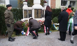 Eine Gruppe von Menschen versammelt sich um ein Denkmal. Ein Mann in Uniform legt Blumen nieder, während andere zusehen.
