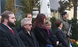 Eine Gruppe von Menschen steht draußen in der Nähe eines Gebäudes. Ein Mann in Uniform hält ein Gewehr. Ein Mann trägt eine rote Krawatte.