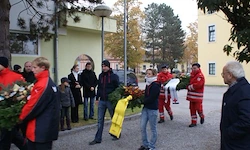 Eine Gruppe von Menschen steht mit Blumen vor einem Gebäude. Ein Mann in einem roten Jackenmantel geht mit Blumen weg. Bäume und gelbe Gebäude im Hintergrund.