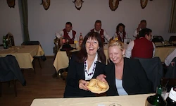 Zwei Frauen sitzen an einem Tisch in einem Restaurant, lächeln und eine hält einen Teller mit Brot. Hinter ihnen sitzen mehrere Personen an Tischen, einige tragen rote Westen.