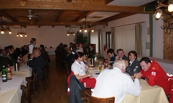 Ein Restaurant mit mehreren Tischen und Stühlen, einer hölzernen Decke und Kronleuchtern. Menschen sitzen an den Tischen, einige stehen. Der Raum ist gut beleuchtet.