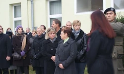 Eine Gruppe von Menschen steht vor einem Gebäude, einige in Uniform, andere in Mänteln. Sie scheinen auf etwas zu warten oder zu beobachten.