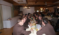 Mehrere Männer in Anzügen sitzen an langen Tischen in einem Speisesaal, führen Gespräche und trinken aus Gläsern. Der Raum hat Holzböden und dekorative Kronleuchter.