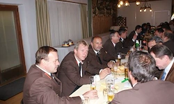 Eine Gruppe von Männern in formellen Anzügen sitzt um einen langen Tisch herum, trinkt Bier und führt Gespräche. Der Raum ist gut beleuchtet mit mehreren Lampen.