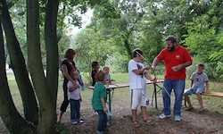 Eine Gruppe von Menschen, einschließlich Kindern, steht um einen Tisch in einem Park herum, wobei ein Mann eine Aktivität mit einem Bogen demonstriert.