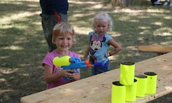 Zwei kleine Mädchen spielen mit Wasserpistolen und gelben Eimern auf einer Picknick-Bank im Park.