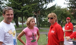 Drei Personen, zwei Frauen und ein Mann, stehen in einem Park. Eine Frau trägt eine Halskette, die andere ein rotes Shirt. Hinter ihnen stehen zwei Männer in roten Shirts bei einem Tisch.