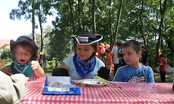 Drei Kinder sitzen an einem Picknicktisch, eines trägt einen Cowboyhut und eine Bandana. Um sie herum stehen Bäume und Menschen im Hintergrund.
