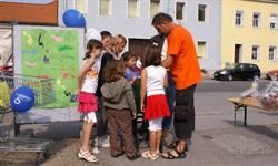 Eine Gruppe von Kindern und einem Erwachsenen stehen draußen. Sie sind vor einem Tisch mit Luftballons und einem gelben Gebäude im Hintergrund.