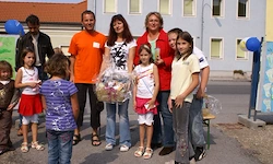 Eine Gruppe von Kindern und Erwachsenen steht vor einem Gebäude und posiert für ein Foto. Sie halten Luftballons und Geschenke. Die Kinder tragen bunte Kleidung und Sandalen.