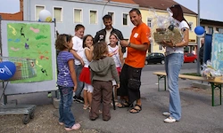 Eine Gruppe von Kindern und Erwachsenen steht um einen Tisch herum, ein Mann in Orange gibt ihnen Geschenke und eine Frau in Weiß hält einen Korb. Im Hintergrund sind Autos und Gebäude zu sehen.