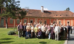 Eine große Gruppe von Menschen steht auf dem Rasen vor einem historischen, rotgedeckten Gebäude mit mehreren Fenstern.
