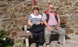 Ein älteres Paar sitzt auf einer Holzbank vor einer Steinmauer. Die Frau hält eine schwarze Tasche. Der Mann trägt eine Brille und eine Uhr.