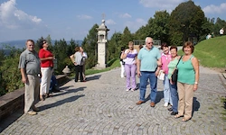Eine Gruppe von Menschen steht auf einem Kopfsteinpflasterweg unter blauem Himmel, mit Bäumen und einem Denkmal im Hintergrund.