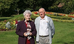 Ein älteres Paar steht in einem Garten und lächelt für ein Foto. Die Frau hält einen Preis. Dahinter bilden farbenfrohe Blumen und Bäume eine üppige Kulisse.