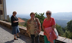 Drei Frauen stehen auf einem gepflasterten Weg, eine in einem schwarzen Shirt und einer Jeansshorts. Die anderen beiden tragen blumige und rosa Shirts, Sonnenbrillen und halten Taschen. Hinter ihnen befindet sich eine Mauer, Bäume und Berge.