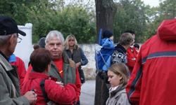 Eine Gruppe von Menschen in roten Jacken und Mützen steht in einem Außenbereich, möglicherweise an einem sonnigen Tag. Bäume und eine weiße Mauer sind im Hintergrund sichtbar.
