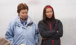 Zwei Frauen stehen nebeneinander. Eine trägt eine blaue Jacke, die andere eine schwarze und rote Jacke mit einem roten Stirnband. Beide haben ernste Gesichtsausdrücke.