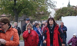 Eine Gruppe von Menschen, darunter Frauen in roten Jacken, steht an einem sonnigen Tag im Freien. Eine Frau spricht in ein Mikrofon, während Bäume und Gebäude im Hintergrund zu sehen sind.