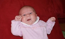 Ein Babymädchen in einem rosa Outfit liegt auf einer roten Decke, lächelt und hebt beide Arme.