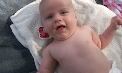 Ein Baby liegt auf einem Handtuch, trägt ein Windel und ein Schnuller liegt neben ihm auf dem Handtuch.