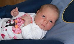 Ein Neugeborenes in einem weißen Langarmshirt mit Blumenmustern liegt auf einem blauen Kissen. Das Baby hat blaue Augen und schaut mit einem leichten Lächeln nach vorne.