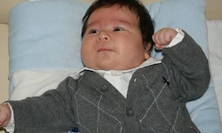 Ein Baby liegt mit erhobenen Armen auf einem blauen Kissen und trägt einen grauen Pullover mit Rautenmuster.