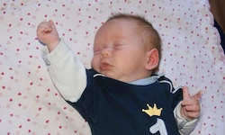 Ein schlafendes Baby in einem dunkelblauen Shirt mit der Nummer eins darauf, auf einer weißen Decke mit roten Herzen liegend.