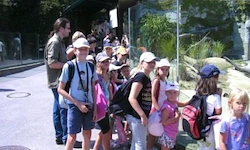 Eine Gruppe von Menschen, darunter Kinder und Erwachsene, warten in einer Schlange, wahrscheinlich in einem Zoo, tragen Hüte und haben Rucksäcke.