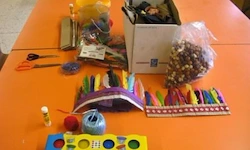 Ein orangefarbener Tisch ist mit verschiedenen Bastelutensilien gefüllt, darunter Scheren, bunte Stifte, eine Spule Garn und eine Schachtel mit Perlen. Ein farbenfroher Kopfschmuck und andere dekorative Gegenstände sind ebenfalls vorhanden.
