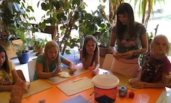 Eine Gruppe von Frauen und Kindern sitzt um einen Tisch herum und ist mit Bastelarbeiten beschäftigt. Sie sind von Topfpflanzen und einem Gebäude im Hintergrund umgeben.