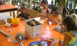 Eine Gruppe von Kindern sitzt um einen orangefarbenen Tisch herum und beschäftigt sich mit einer Bastelaktivität. Sie haben verschiedene Bastelmaterialien wie eine Schachtel, Plastikbehälter und bunte Materialien.