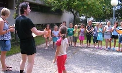 Ein Mann in schwarzer Kleidung unterrichtet eine Gruppe von Kindern einen Tanz im Freien. Hinter ihnen steht ein Gebäude mit Bäumen.