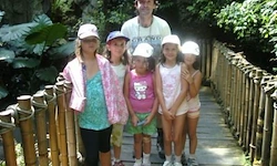 Eine Gruppe von Kindern und ein Mann stehen auf einer Holzbrücke im Wald. Der Mann trägt eine Halskette und ein weißes Hemd.