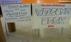 Zwei Schilder auf einem Schreibtisch, eines mit bunten Buchstaben 'Welcome Desk SPO' und ein weiteres mit blauem Text 'Herzliche Einladung 30. Ferien zu Hause ist auf Ausflug'.