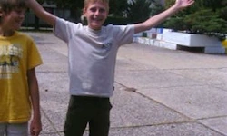 Zwei Jungen stehen draußen. Ein Junge hat die Arme erhoben und lächelt. Der andere Junge trägt ein gelbes T-Shirt und schaut in die Kamera. Dahinter befindet sich ein Beton-Gehweg mit Pflanzen.