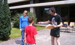 Ein Mann in einem schwarzen T-Shirt liest einem Jungen in einem roten T-Shirt etwas vor. Eine Frau in einem blauen T-Shirt steht in der Nähe. Sie stehen vor einem Gebäude mit Glasfenstern.