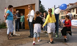 Eine Gruppe von Menschen steht draußen in der Nähe eines Gebäudes mit Luftballons, darunter eine Frau auf einem Hocker. Einige tragen Sandalen und Turnschuhe.