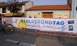 Ein Banner, das das Radrekordtag-Fahrradevent bewirbt, ist auf einer orangefarbenen Barriere mit einem geparkten Fahrrad zu sehen. Der Banner trägt den Text 'Radrekordtag' und ermutigt zum Radfahren für den Klimaschutz.