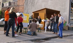 Eine Gruppe von Menschen steht um einen Stand auf dem Bürgersteig, ein Mann kniet und arbeitet an einem Projekt. Hinter ihnen steht eine kleine Struktur und ein Schild, und ein Auto ist in der Nähe geparkt.