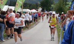 Marathonläufer joggen auf einer Straße, mit Zuschauern auf beiden Seiten. Im Hintergrund sind mehrere Zelte und Bäume zu sehen.