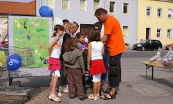 Eine Gruppe von Kindern und Erwachsenen steht um einen Mann in einem orangefarbenen Shirt herum und betrachtet ein Plakat mit Luftballons vor einem gelben Gebäude im Hintergrund.