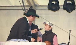 Ein Mann mit schwarzem Hut hält ein Mikrofon und interviewt einen jungen Jungen in einer weißen Mütze und einem rosa Shirt, der vor einem Tisch steht.