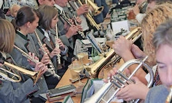 Eine Gruppe von Musikern in Uniformen spielt ihre Trompeten und Flöten bei einem Konzert, mit einem verschwommenen Publikum im Hintergrund.
