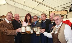 Eine Gruppe von Menschen, einschließlich Männer und Frauen, halten Bierkrüge unter einem Zelt, möglicherweise auf einem Bierfestival. Sie tragen alle formelle Kleidung, einige von ihnen tragen Krawatten.