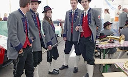 Fünf Personen in traditioneller Kleidung stehen in einem Zelt und halten Musikinstrumente. Ein Tisch mit rot-weiß kariertem Tischtuch befindet sich in der Nähe.