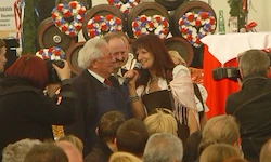 Ein Mann und eine Frau sprechen in ein Mikrofon bei einer Veranstaltung, mit einer großen Menschenmenge um sie herum und Kränzen im Hintergrund.