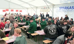 Eine große Gruppe von Menschen in grünen Uniformen und Lederjacken sitzt an Tischen in einem großen Zelt mit einem Banner, das 'SPARKASSE Weinsburg-Druck' liest.