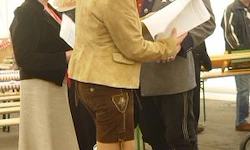 Eine Frau in einer beigen Jacke und braunen Shorts hält ein Papier in der Hand und steht zwischen zwei Männern in traditioneller Kleidung, möglicherweise bei einer Feier.
